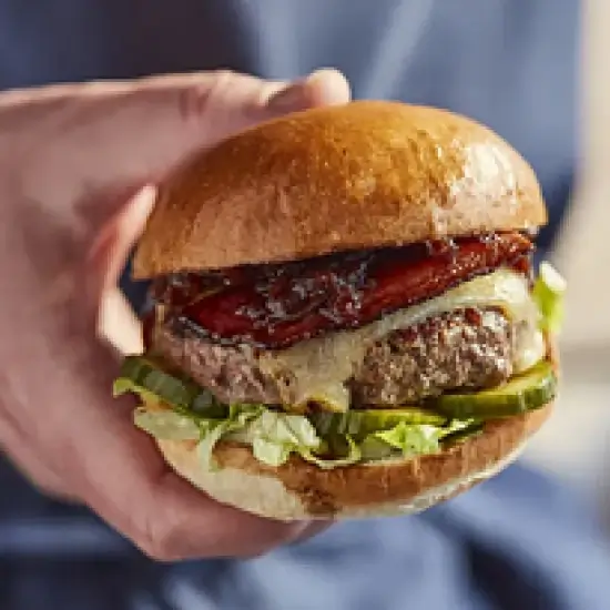 Hand holding an Honest beef burger with lettuce, pickles, melted cheese, and roasted red peppers in a brioche bun.